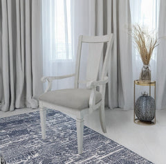 A white wooden dining armchair with a light beige upholstered seat cushion, positioned on a blue and white patterned rug in a bright room with gray curtains.