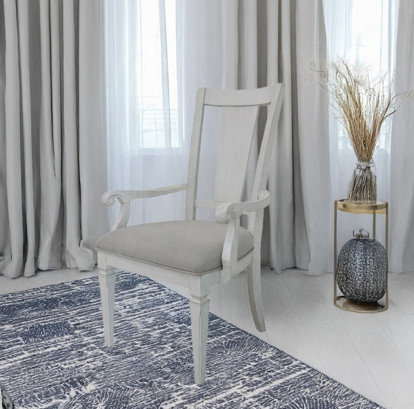 A white wooden dining armchair with a light beige upholstered seat cushion, positioned on a blue and white patterned rug in a bright room with gray curtains.