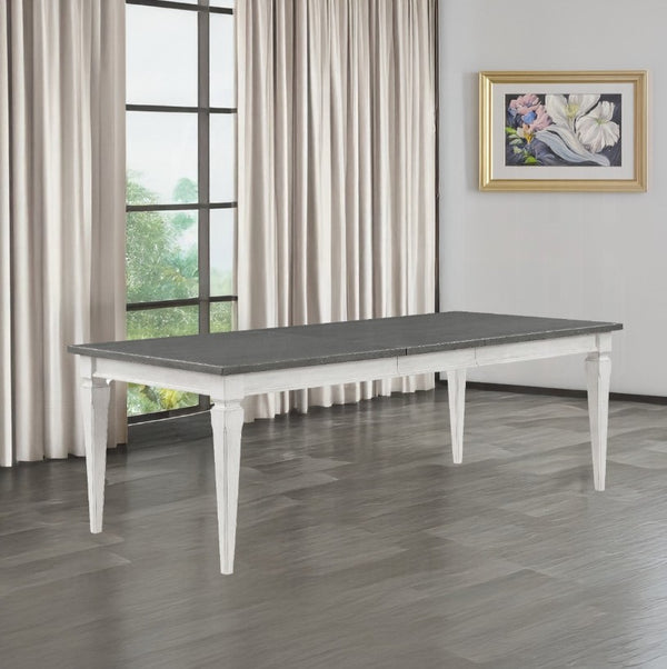 A rectangular dining table with a dark gray stone-look top and white legs, shown in a staged room with beige curtains, a window, and a framed floral print on the wall.
