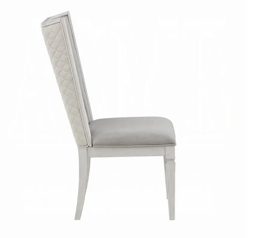 A white dining chair with a light gray upholstered seat and backrest, positioned on a gray and white patterned rug in a bright room with sheer curtains and windows.