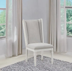 A white dining chair with a light gray upholstered seat and backrest, positioned on a gray and white patterned rug in a bright room with sheer curtains and windows.