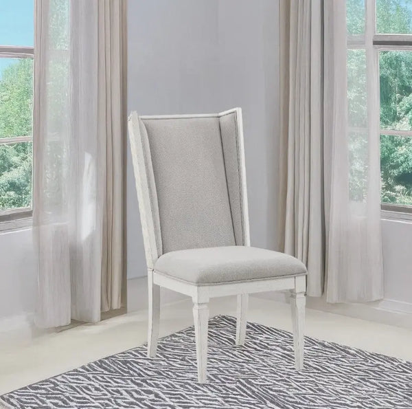 A white dining chair with a light gray upholstered seat and backrest, positioned on a gray and white patterned rug in a bright room with sheer curtains and windows.