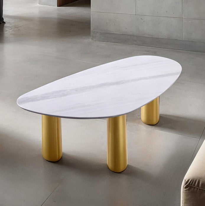 Naynem Coffee Table Oval Sintered Stone White Gold Finish An oval coffee table with a white marble-look top and four cylindrical gold legs, shown in a modern interior setting with a concrete floor and gray tiled wall.