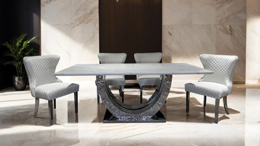 Primenova Horseshoe Shaped Sintered Stone Chrome White Dining Table A modern dining room set featuring a rectangular white marble-top table with a sculptural dark gray stone base, surrounded by four black upholstered dining chairs with silver metal legs, set in a luxury interior with marble floors and walls.