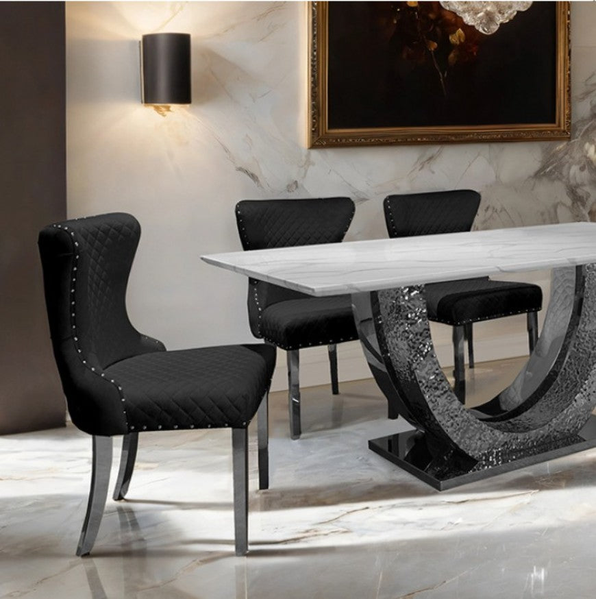 Primenova Horseshoe Shaped Sintered Stone Chrome White Dining Table A modern dining room set featuring a rectangular white marble-top table with a sculptural dark gray stone base, surrounded by four black upholstered dining chairs with silver metal legs, set in a luxury interior with marble floors and walls.
