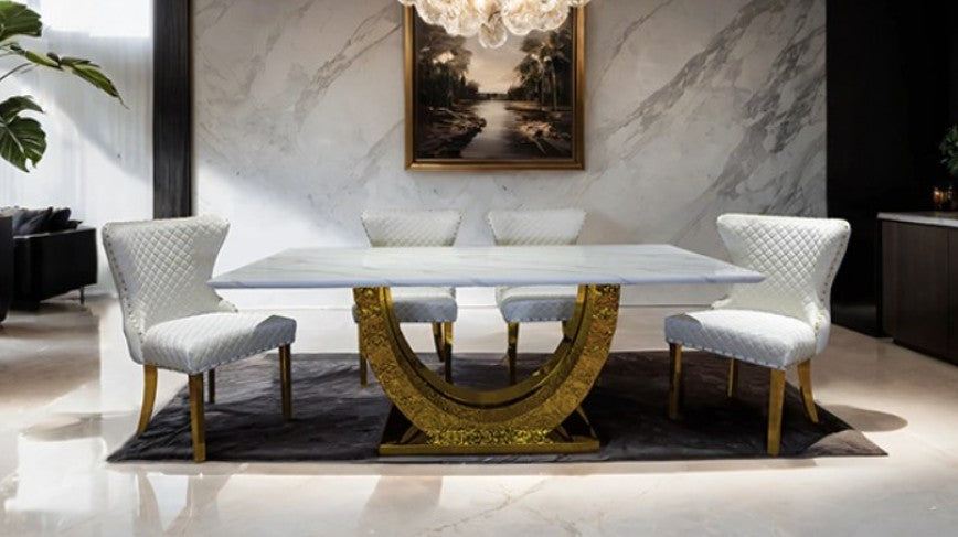 Primenova Horseshoe Shaped Sintered Stone Gold White Dining Table A modern dining room scene featuring a rectangular white dining table with a sculptural gold curved pedestal base, surrounded by four black upholstered dining chairs with gold legs, set in a contemporary interior with a fireplace and marble floor.