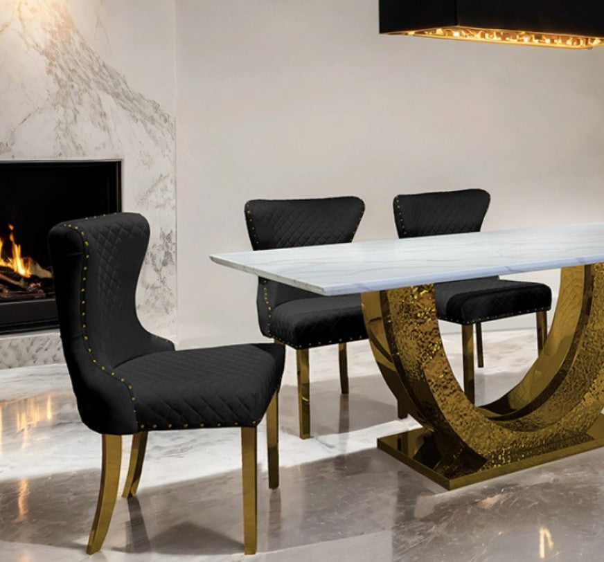 Primenova Horseshoe Shaped Sintered Stone Gold White Dining Table A modern dining room scene featuring a rectangular white dining table with a sculptural gold curved pedestal base, surrounded by four black upholstered dining chairs with gold legs, set in a contemporary interior with a fireplace and marble floor.