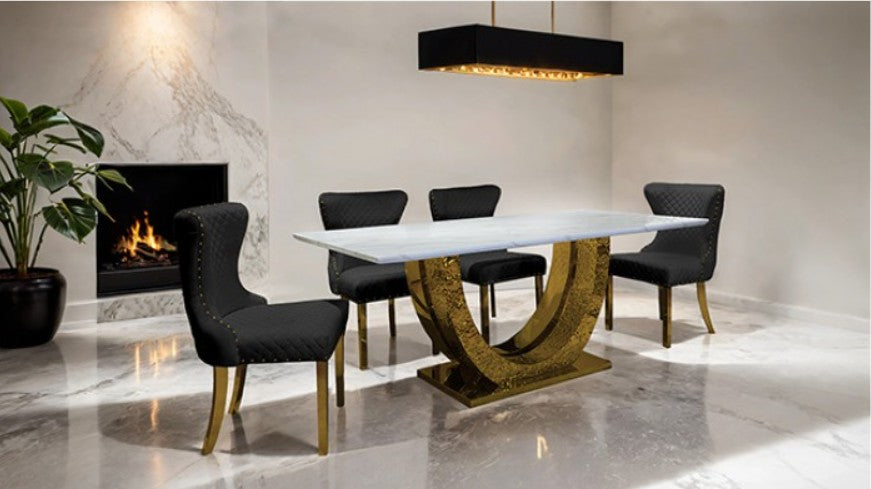 Primenova Horseshoe Shaped Sintered Stone Gold White Dining Table A modern dining room scene featuring a rectangular white dining table with a sculptural gold curved pedestal base, surrounded by four black upholstered dining chairs with gold legs, set in a contemporary interior with a fireplace and marble floor.