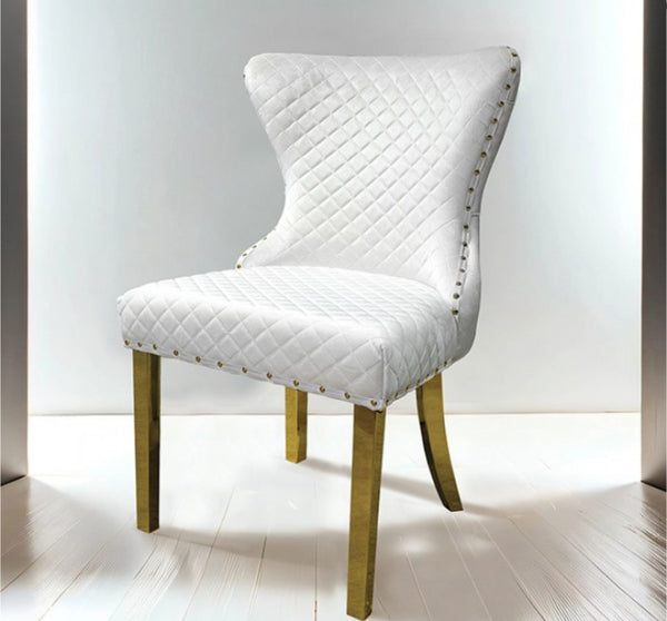 Primenova Upholstered Gold White Dining Chair A white quilted upholstered dining chair with gold-toned metal legs and nailhead trim, shown in a bright studio setting with a white wall and light wood floor.