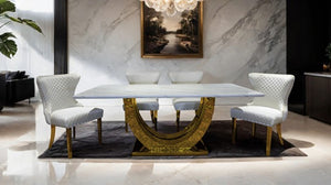 Primenova Upholstered Gold White Dining Chair A luxury dining table set featuring a rectangular white marble tabletop on an ornate gold crescent-shaped pedestal base, surrounded by four white quilted velvet dining chairs with gold legs, set on a dark rug in a modern dining room.
