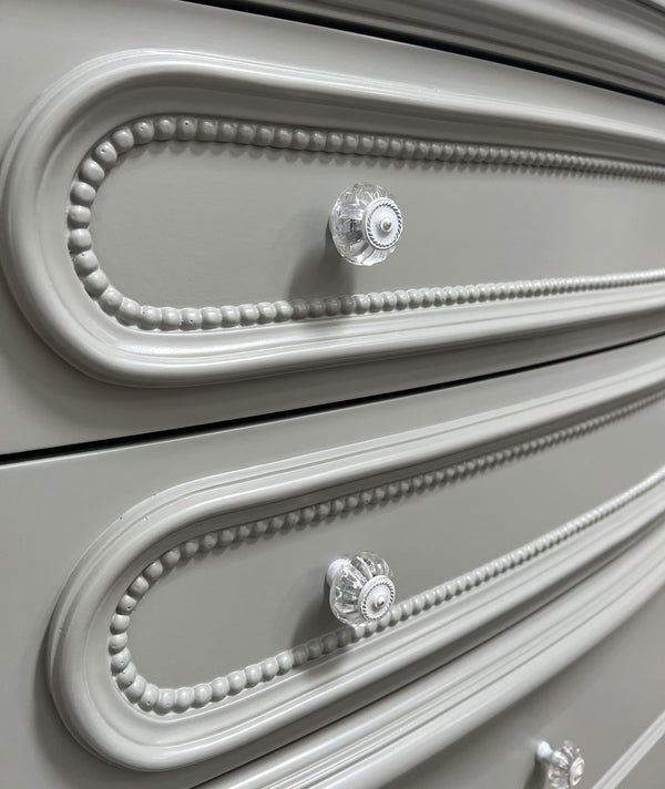 Primrose Gray Chest with Floral Accent Close-up of a light gray wooden dresser with ornate raised drawer fronts, beaded trim details, and clear faceted crystal-style drawer knobs.