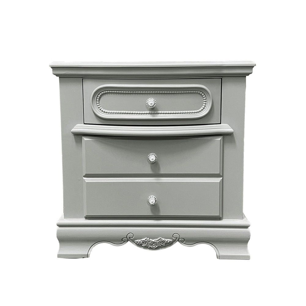 Primrose Gray Nightstand with Floral Accent A light gray three-drawer nightstand in a bedroom setting, styled with a table lamp, a small globe, and a cup on top, next to a matching gray bed frame.