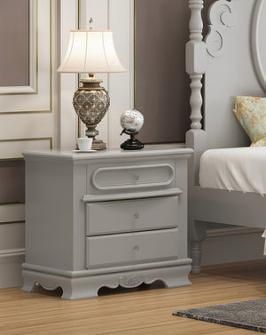 Primrose Gray Nightstand with Floral Accent A light gray three-drawer nightstand in a bedroom setting, styled with a table lamp, a small globe, and a cup on top, next to a matching gray bed frame.