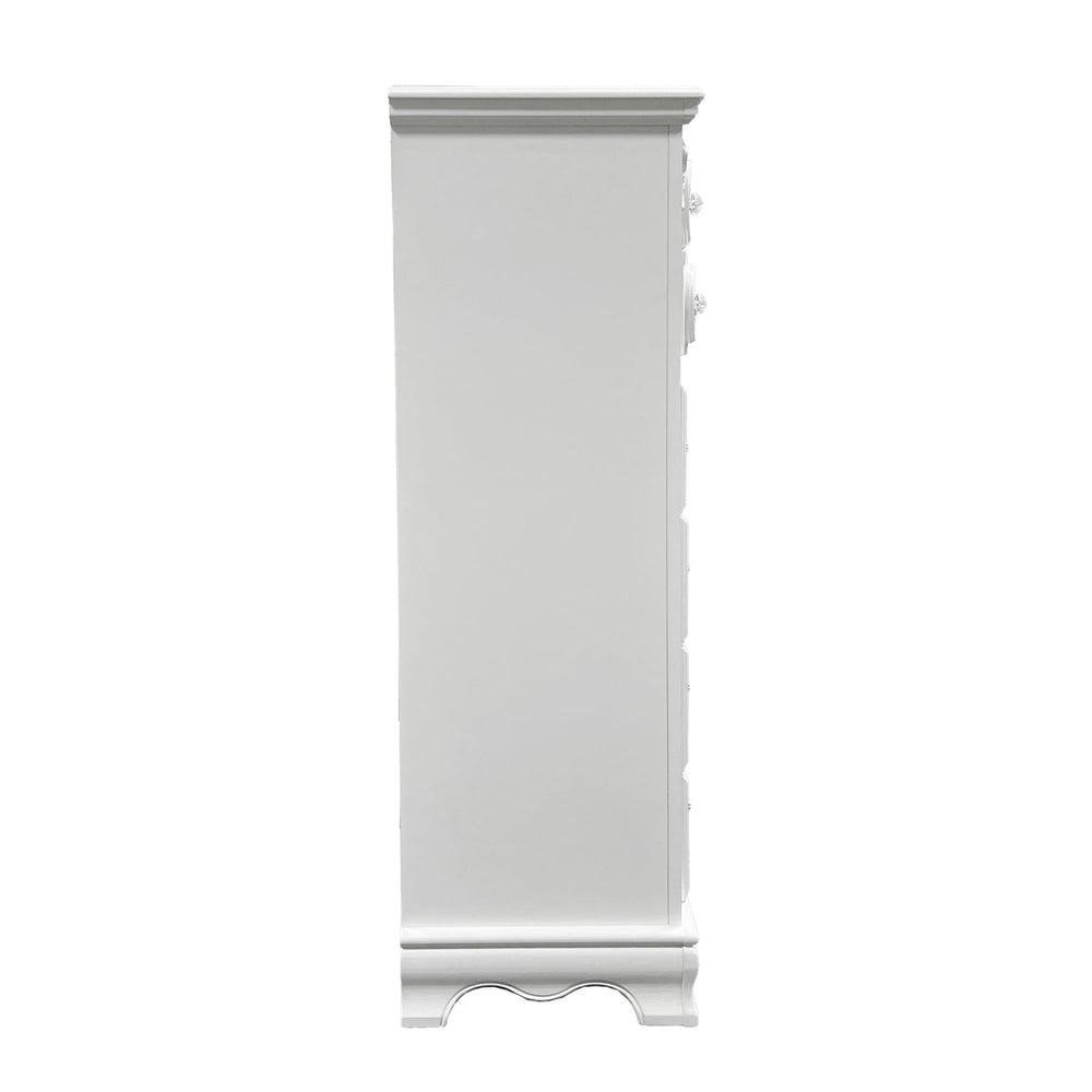 Primrose White Chest with Floral Accent A tall white wooden five-drawer chest of drawers with ornate silver handles and decorative drawer fronts, shown in a bright room with sheer white curtains, gray drapes, a brown floor, and a light rug. A small potted plant sits on top.