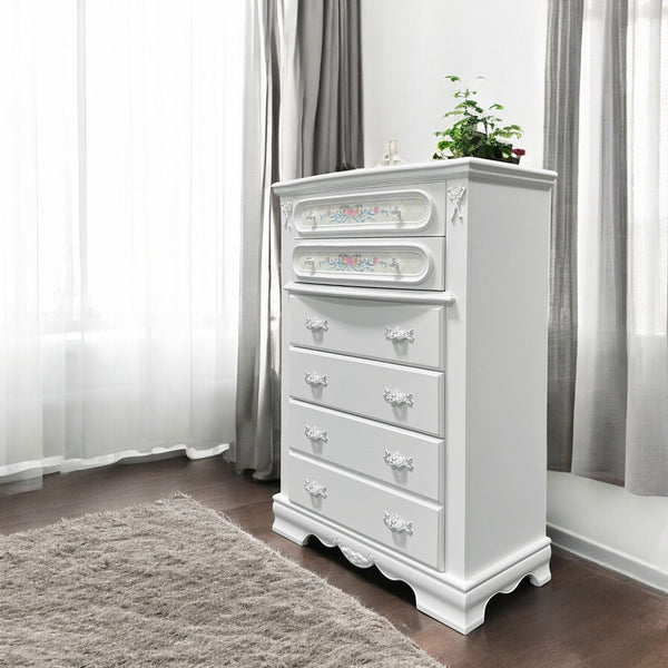Primrose White Chest with Floral Accent A tall white wooden five-drawer chest of drawers with ornate silver handles and decorative drawer fronts, shown in a bright room with sheer white curtains, gray drapes, a brown floor, and a light rug. A small potted plant sits on top.