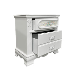 Primrose White Nightstand with Floral Accent A white wooden nightstand with three drawers, featuring ornate silver handles, a floral inlay on the top drawer, and carved decorative details on the side.