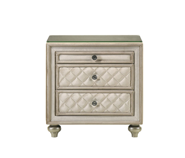Sonya Beige Velvet Upholstered Traditional Style Nightstand A three-drawer nightstand with a distressed beige and silver finish, featuring quilted cream leatherette drawer fronts and a clear glass top, set against a plain white background.