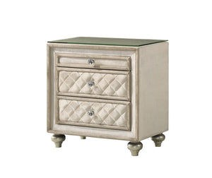 Sonya Beige Velvet Upholstered Traditional Style Nightstand A three-drawer nightstand with a distressed cream and silver finish, featuring quilted diamond-patterned drawer fronts, round silver knobs, and a clear glass top, shown on a plain white background.