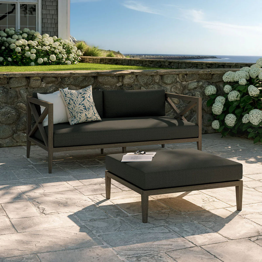 Sunshore Flint Gray Loveseat and Ottoman Outdoor Set Outdoor patio set with cushions on a stone patio, surrounded by flowers and a scenic view.