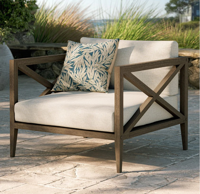 Sunshore Pebble Outdoor Armchair Outdoor chair with wooden frame and beige cushions, featuring a decorative pillow, on a stone patio.