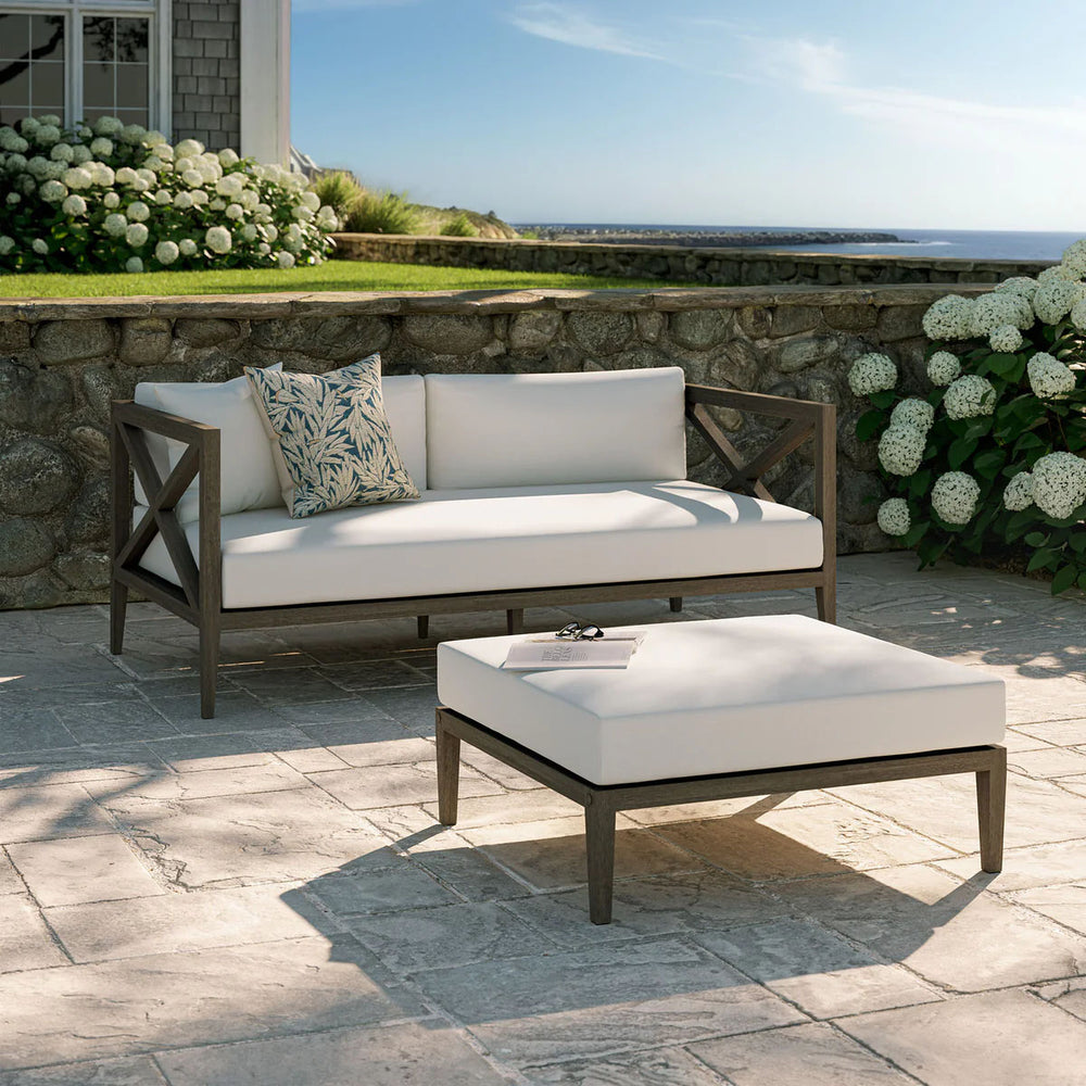 Sunshore Weathered Gray Loveseat and Ottoman Outdoor Set Outdoor patio set with sofa and ottoman on a stone patio, surrounded by flowers and a stone wall.
