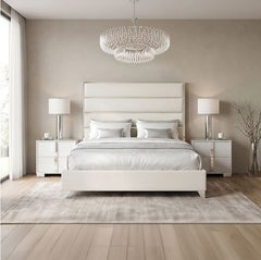 Sysko Glam Silver White Fully Upholstered Bed A modern bedroom featuring a white upholstered bed frame with a padded headboard and matching footboard, styled with white bedding, two matching white nightstands with silver lamps, a crystal chandelier, and a light gray rug on light wood flooring.