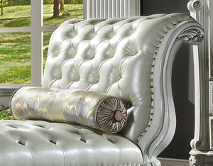 Traditional Style Chaise Bone White Dresden Collection A close-up of an elegant tufted chaise lounge chair in a light silver-gray leather, featuring a matching cylindrical lumbar pillow with a gold and gray damask pattern, set against a dark wall with a window showing a green lawn.