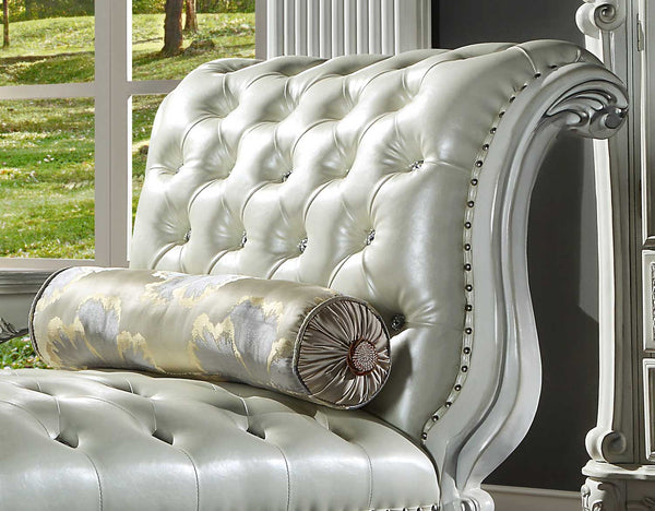 Traditional Style Chaise Bone White Dresden Collection A close-up of an elegant tufted chaise lounge chair in a light silver-gray leather, featuring a matching cylindrical lumbar pillow with a gold and gray damask pattern, set against a dark wall with a window showing a green lawn.