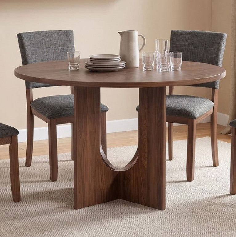 Unberto Walnut Round Dining Table A round wooden dining table with a sculptural pedestal base, surrounded by four gray upholstered dining chairs in a warm, neutral-toned dining room setting.