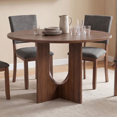 Unberto Walnut Round Dining Table A round wooden dining table with a sculptural pedestal base, surrounded by four gray upholstered dining chairs in a warm, neutral-toned dining room setting.