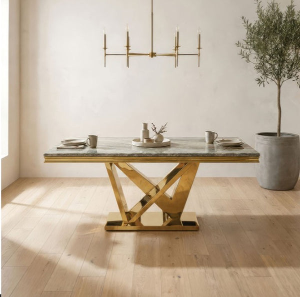 Valdi Gold Gray Sintered Stone Top Dining Table A rectangular dining table with a polished gold metallic base and a gray marble top, styled in a bright, modern dining room with a gold chandelier, light wood floors, and a potted tree.
