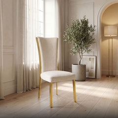Valdi Gold White Upholstered Side Chair (2/CTN) A modern dining chair with a textured cream upholstered seat and back, and light natural wood legs, set in a bright, elegant room with a potted olive tree and warm lighting.