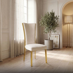 Valdi Gold White Upholstered Side Chair (2/CTN) A modern dining chair with a textured cream upholstered seat and back, and light natural wood legs, set in a bright, elegant room with a potted olive tree and warm lighting.