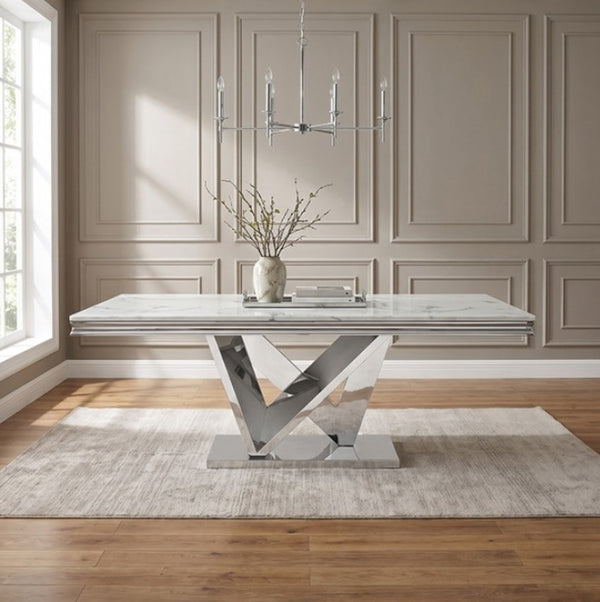 Valdi Silver White Sintered Stone Top Dining Table A modern rectangular dining table with a white marble-look top and a reflective chrome geometric pedestal base, styled in a bright room with beige paneled walls, a light rug, wood floor, and a hanging chandelier.