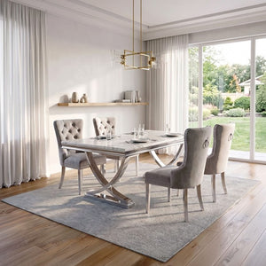 Valdi Silver White Sintered Stone Top V-Style Pedestal Dining Table A modern dining room set featuring a rectangular marble-top dining table with a chrome X-shaped base, surrounded by four tufted gray velvet chairs on a light gray rug. The room has light wood floors, white walls, sheer curtains, and a large window looking out onto a green garden.