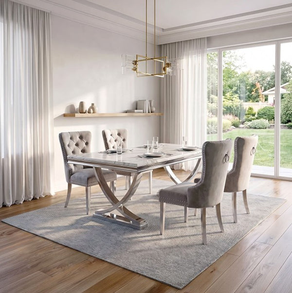 Valdi Silver White Sintered Stone Top V-Style Pedestal Dining Table A modern dining room set featuring a rectangular marble-top dining table with a chrome X-shaped base, surrounded by four tufted gray velvet chairs on a light gray rug. The room has light wood floors, white walls, sheer curtains, and a large window looking out onto a green garden.