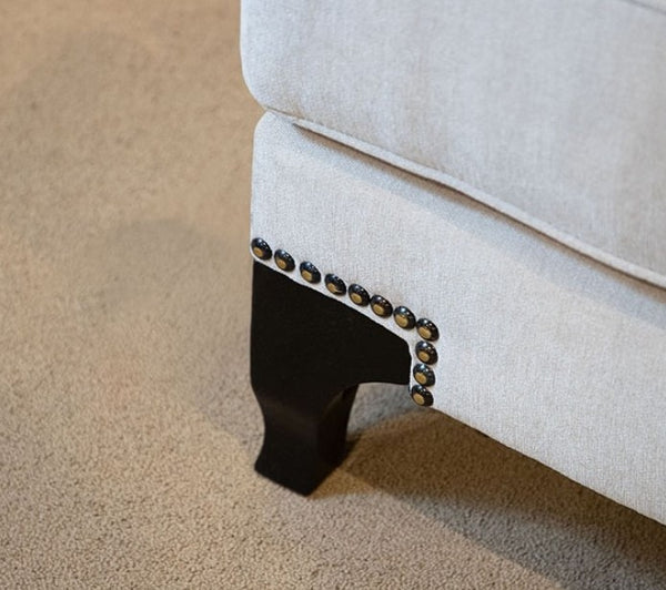 Close-up of a beige upholstered chair with decorative nailhead trim on a carpeted floor.